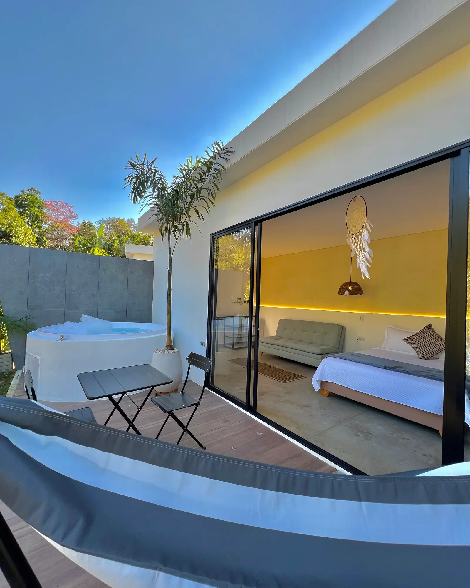 La cabaña doble por fuera: fachada de vidrio, jacuzzi y una palma contra el cielo azul
